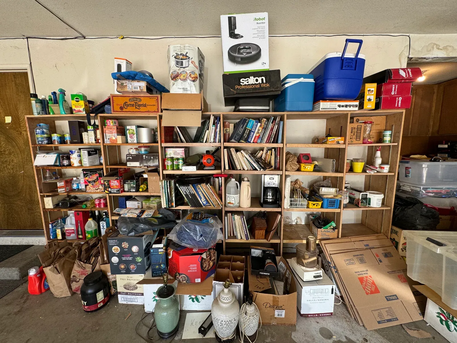 Garage shelving packed with cans, boxes and books, more boxes stacked on top of it, and bags and cartons piled on the floor in front.