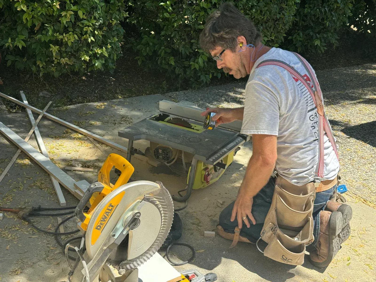 A cabinetmaker kneeling at a table saw and mitre saw set up on a driveway during an install.