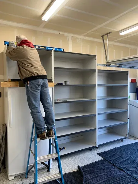 An installer on a step ladder levelling the top of a new cabinet run against a garage wall.
