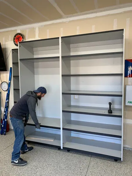 An installer setting the base of two tall open cabinets against a garage wall.